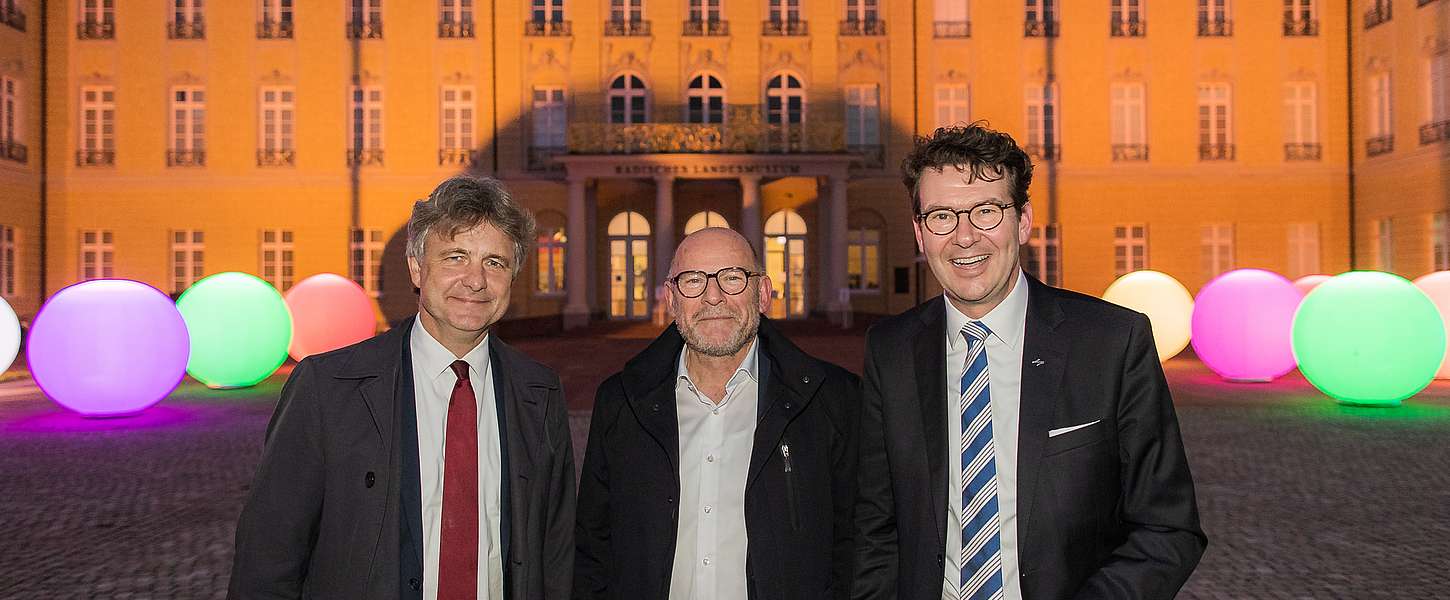 Fünf Männer in Anzügen stehen vor einem KVV-Plakat. Zwei schütteln Hände. Sie halten Urkunden mit dem Wappen Baden-Württembergs in den Händen.