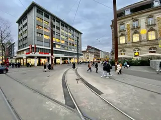 Das Foto zeigt den Gleisbogen am Karlsruher Europaplatz. Menschen überqueren die Kreuzung, im Hintergrund sind eine AVG-Stadtbahn und Häuserfassaden zu sehen.